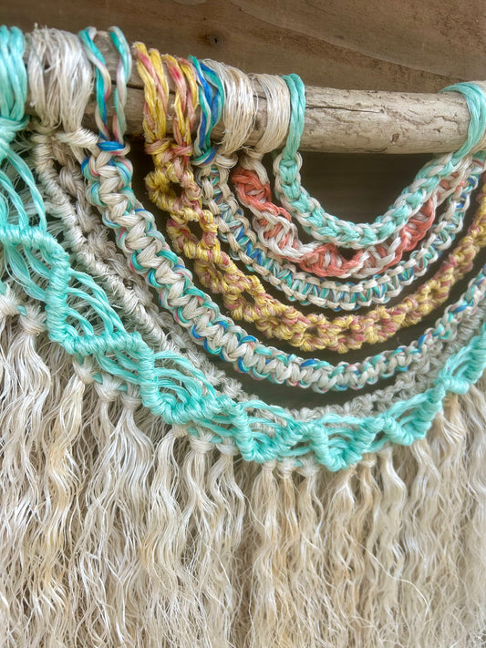 Close up shot of coastal inspired wall hanging handmade from repurposed fishing ropes in soft, sun-bleached tones of green, blue, yellow and coral, with textured weathered white fringe, set on driftwood.