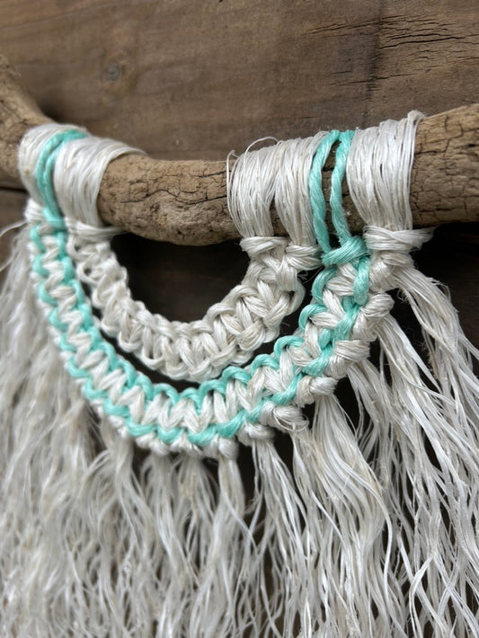 Close-up of reclaimed fishing rope macramé wall hanging in weathered white with a soft green current flowing through, set on driftwood.