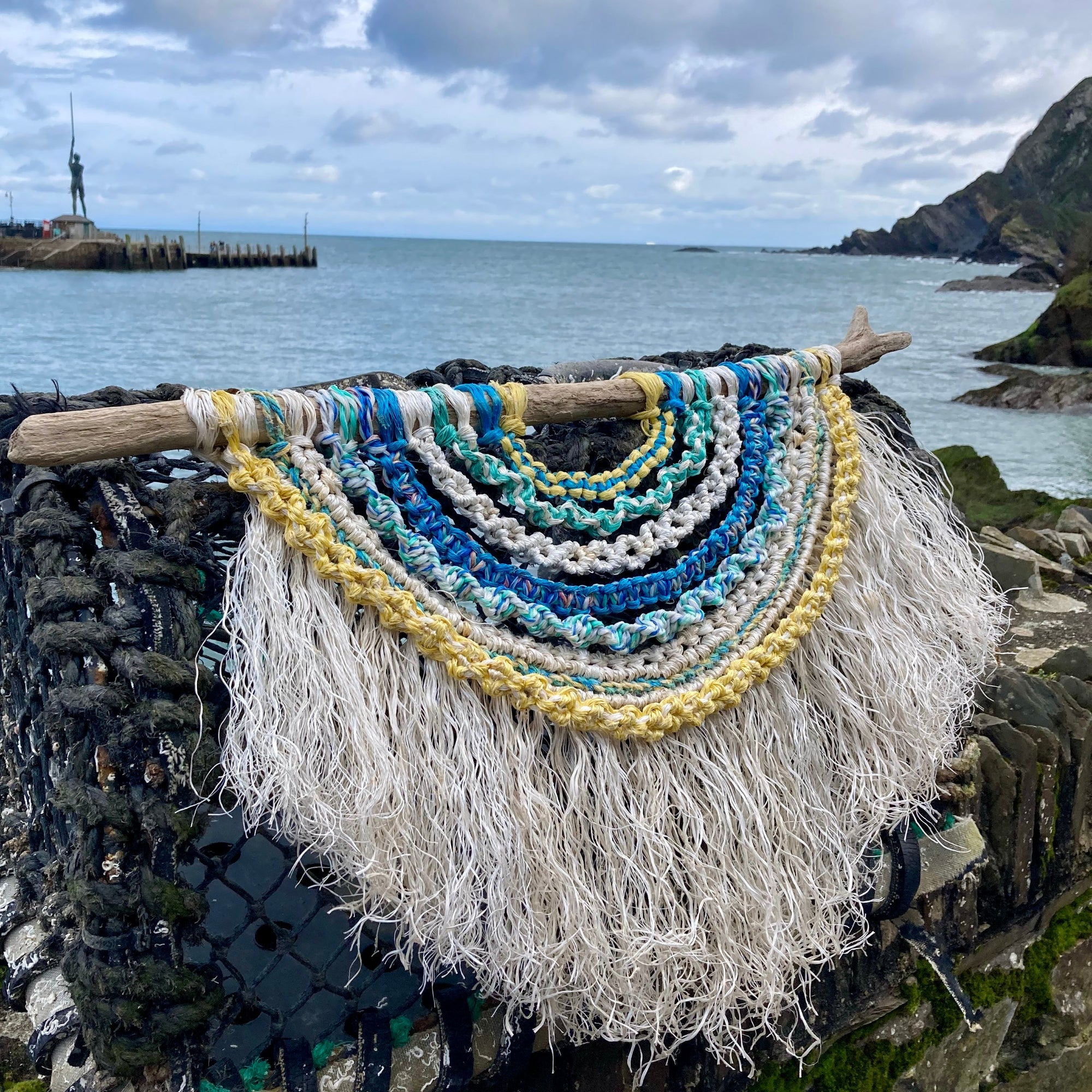 Handmade layered rope wall hanging on driftwood, made from repurposed fishing rope in aged white and coastal tones of sea green, sea blue and sun-softened yellow, photographed on a lobster pot in Ilfracombe Quay overlooking the sea and Damien Hirst's Verity.