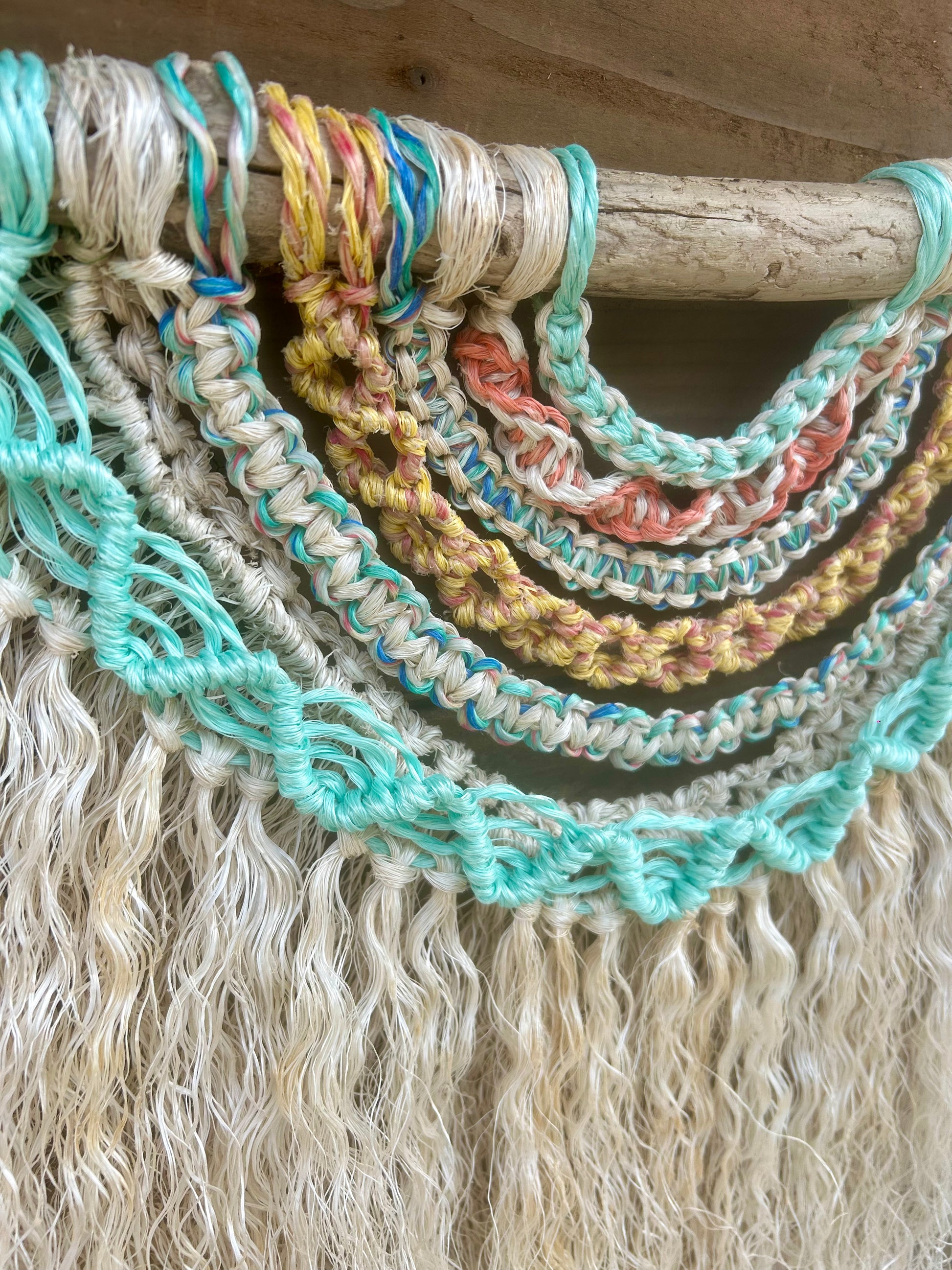 Close up shot of coastal inspired wall hanging handmade from repurposed fishing ropes in soft, sun-bleached tones of green, blue, yellow and coral, with textured weathered white fringe, set on driftwood.