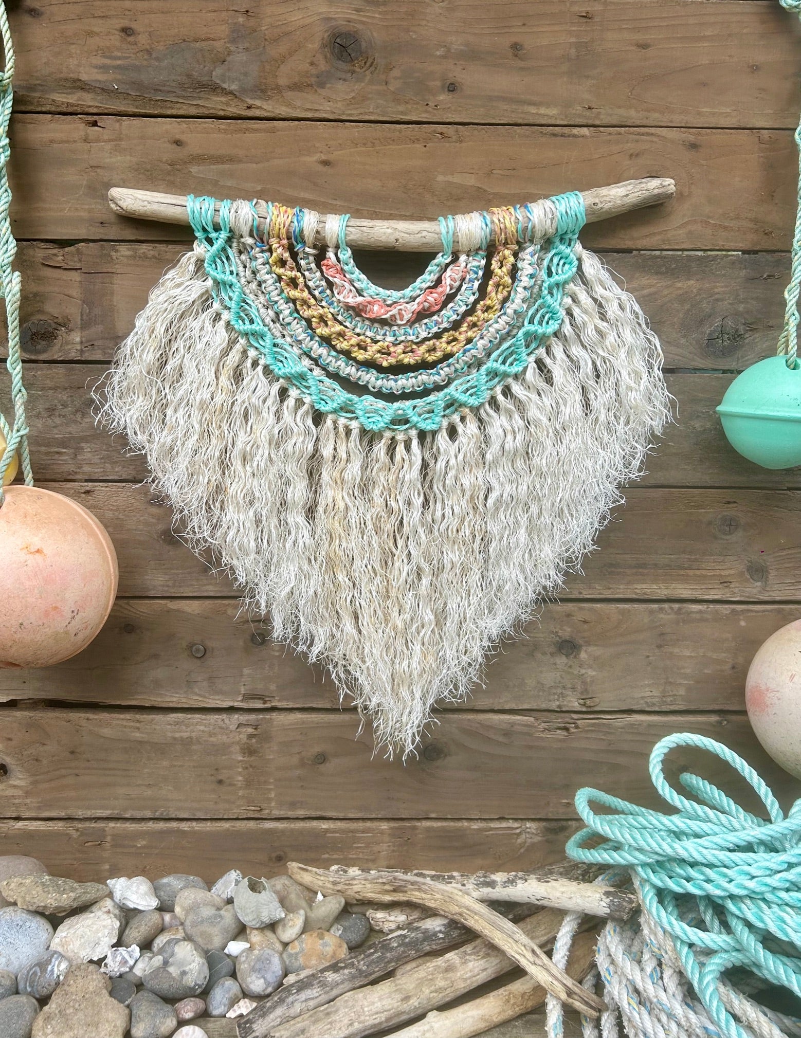 Coastal wall piece sustainably handmade from reclaimed marine ropes. Layered sun-bleached coastal tones of sea blue and green, soft yellow and coral with a weathered white fringe, rust spotted from its previous working life.