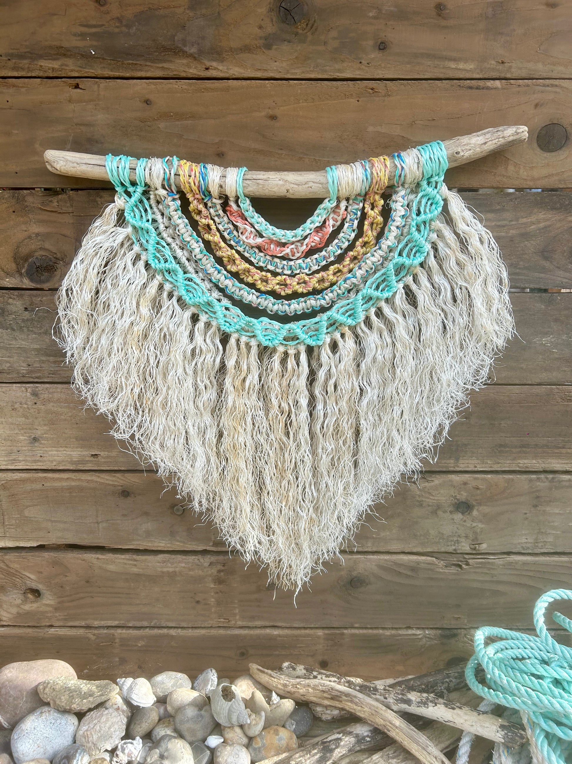 Handmade coastal wall art made from reclaimed fishing ropes. In sun-bleached tones of sea blue and green, soft yellow and coral with a textured white fringe featuring a touch of rust hinting at its working history. Set on driftwood.