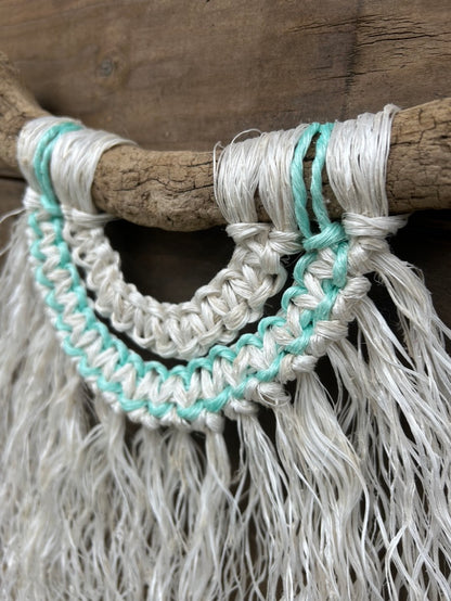 Close-up of reclaimed fishing rope macramé wall hanging in weathered white with a soft green current flowing through, set on driftwood. 