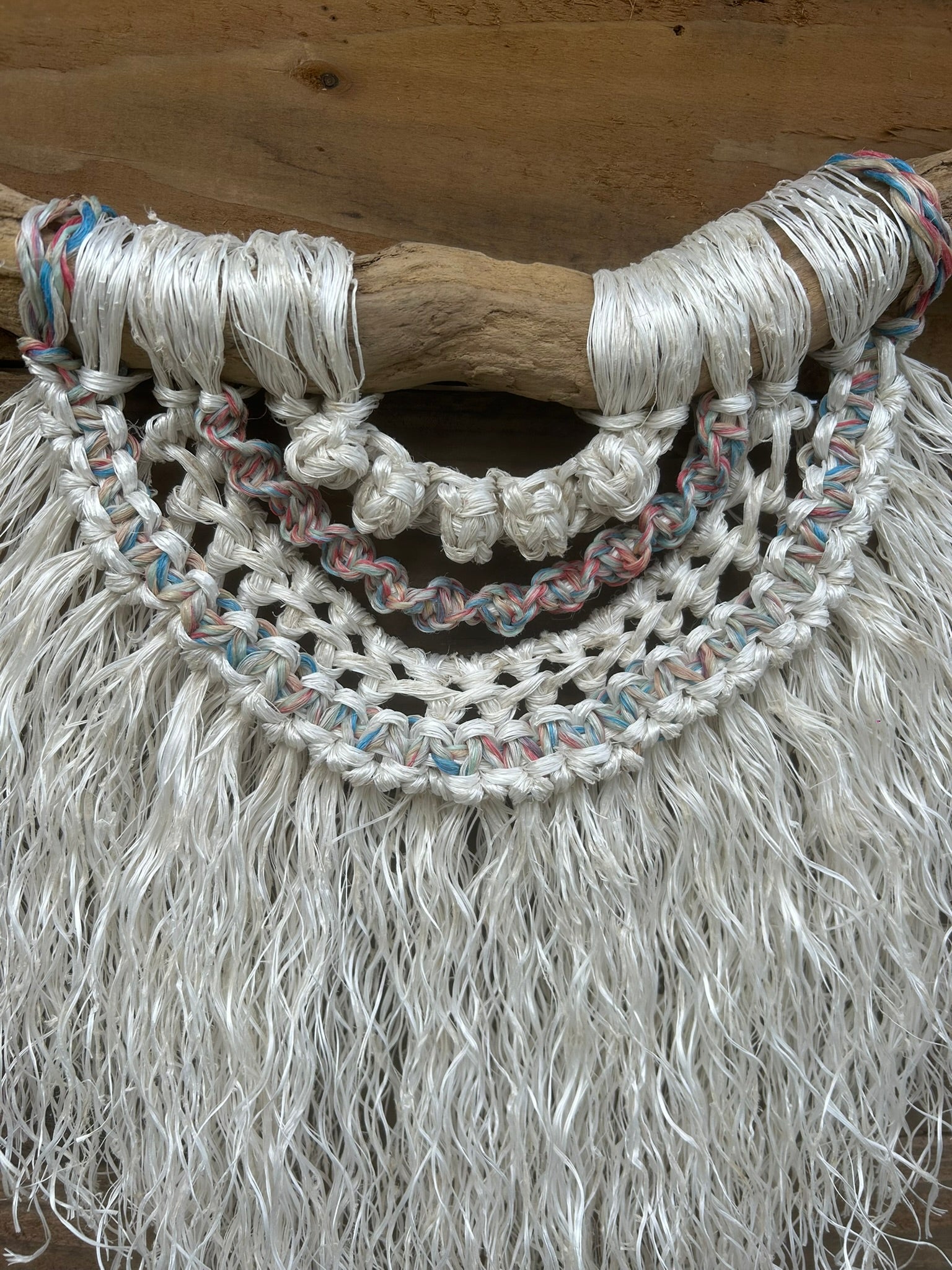 Sustainable coastal wall piece handcrafted from reclaimed fishing rope with a soft white fringe and sun-bleached tones of red, white and blue, set on driftwood.