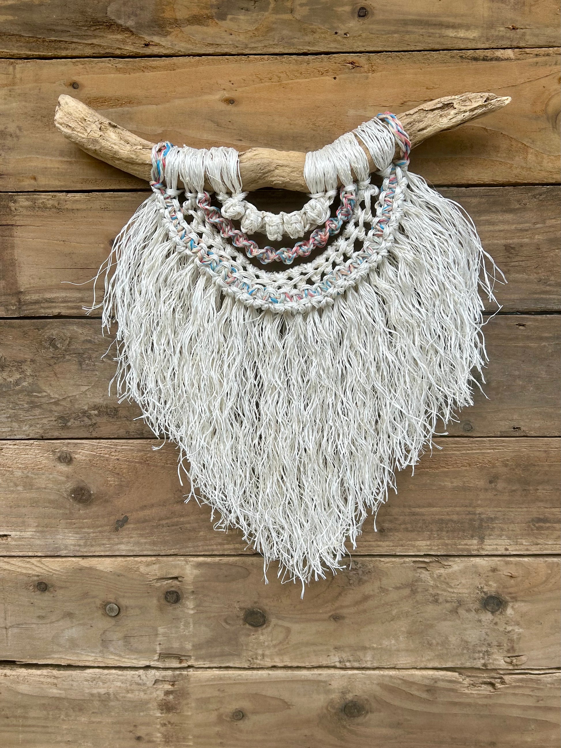 Sustainable rope wall art hand-knotted from repurposed rope with a soft white fringe and sun-bleached tones of red, white and blue, set on driftwood.