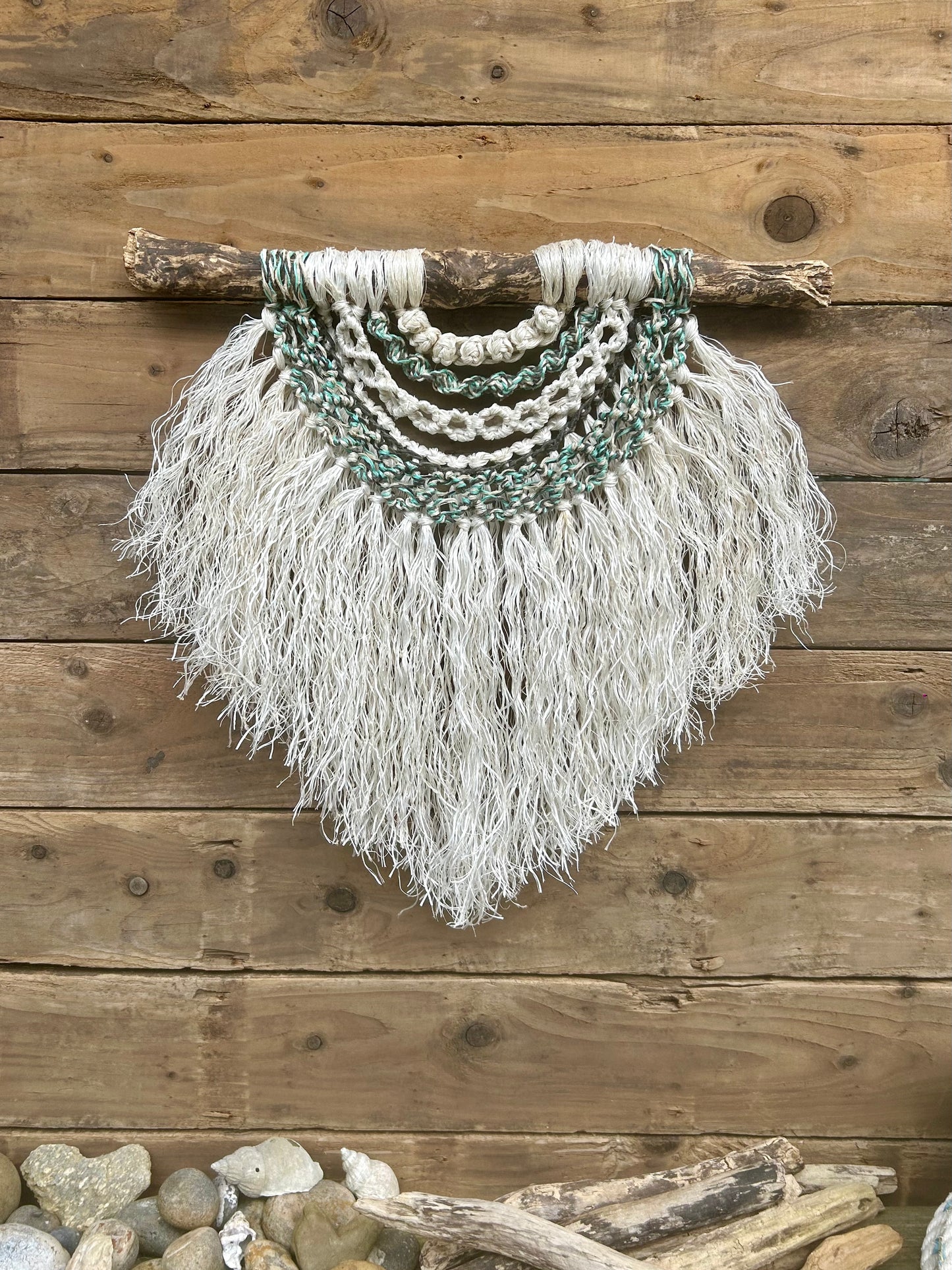 Textile art piece handcrafted from retired fishing ropes, with a weathered white fringe and tidal tones of green and black, set on driftwood.