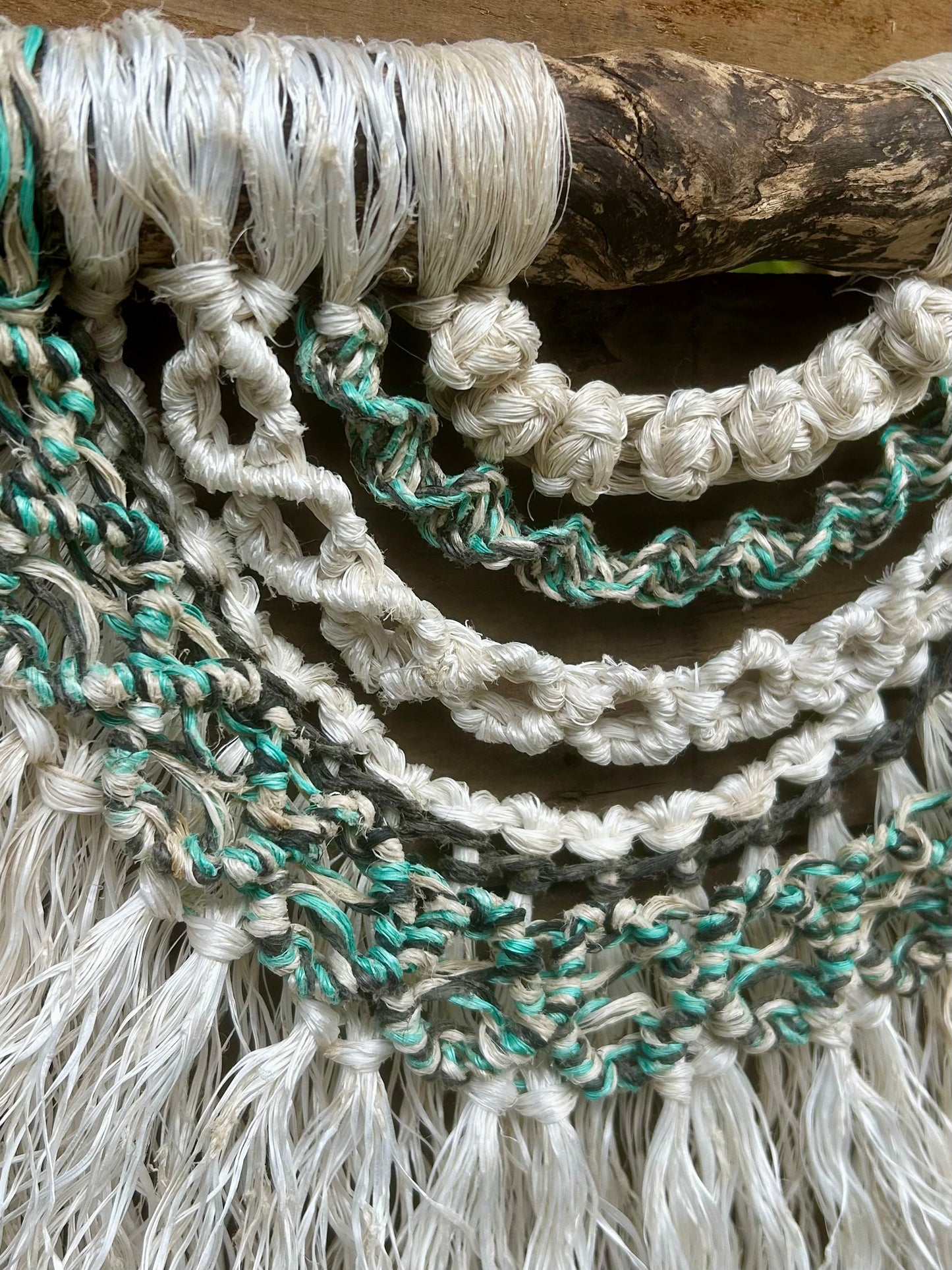 Close-up of coastal inspired macramé wall art, handmade from repurposed fishing ropes in rock pool tones of soft white, green and black, set on driftwood.