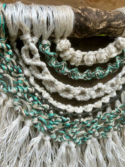 Close-up of coastal inspired macramé wall art, handmade from repurposed fishing ropes in rock pool tones of soft white, green and black, set on driftwood.