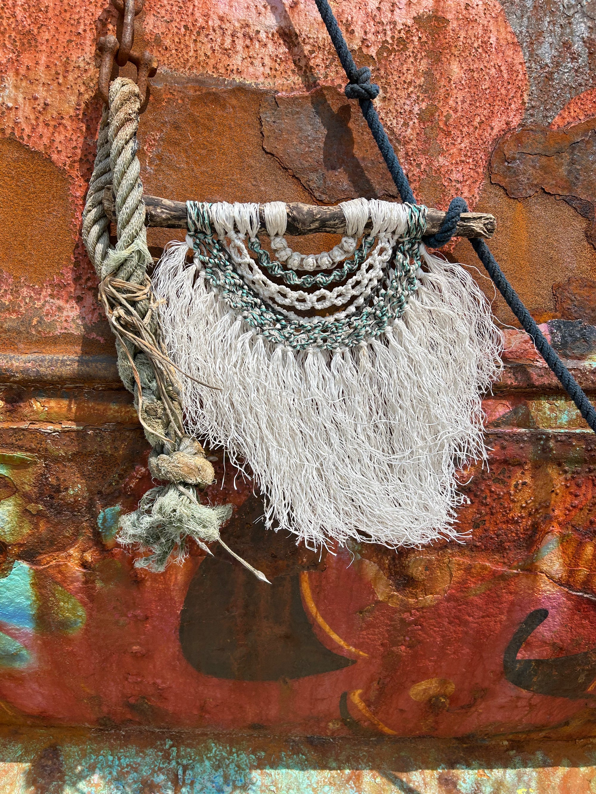 Sustainable coastal wall art made from driftwood and repurposed fishing rope in tidal tones of weathered white, green and black. Photographed on rusted boat in Yelland, North Devon.