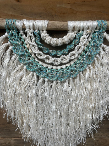 Ocean inspired coastal wall hanging, handcrafted from reclaimed fishing ropes, with a soft white fringe and sea greens and blues, set on driftwood.
