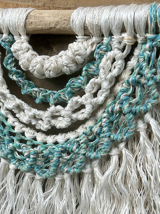 Close-up shot of coastal wall hanging in soft white and sea greens and sea blues, handcrafted from ocean waste, set on driftwood.