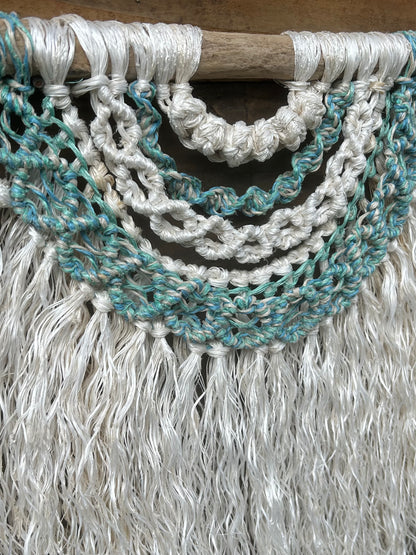 Side shot of macramé wall hanging in coastal tones of sea blues and greens with  a soft white fringe, sustainably made from retired fishing ropes, set on driftwood.