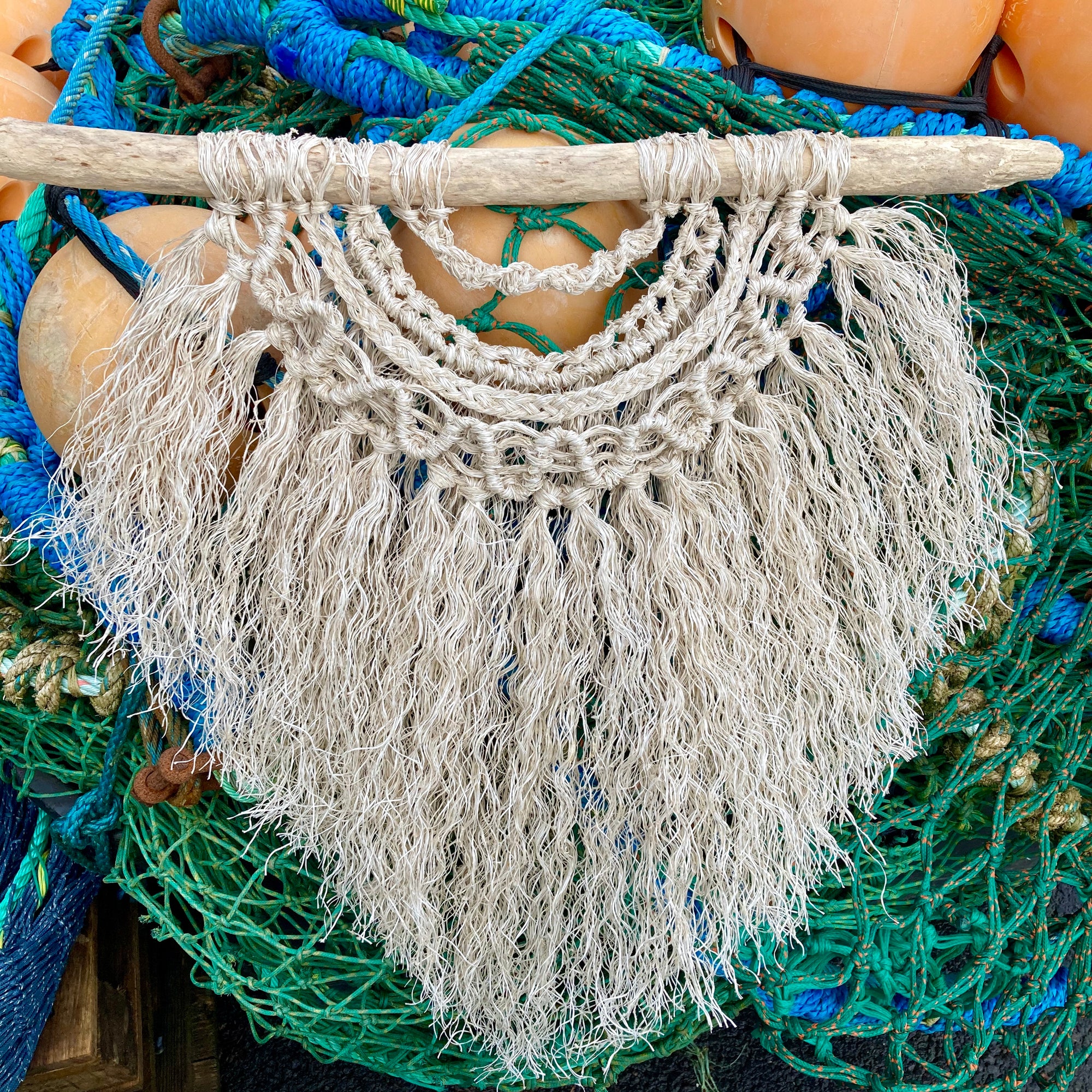 Handcrafted layered coastal wall hanging made from driftwood and repurposed fishing rope in a textured old white tone. Photographed on a pile of fishing nets and buoys in Ilfracombe Quay.