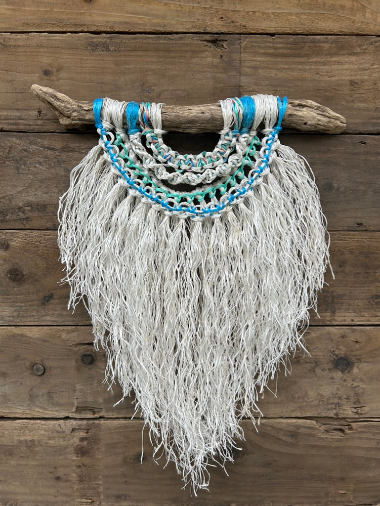 Sustainable macrame wall hanging created from reclaimed fishing rope, in soft white with sea greens and blues, set on driftwood.