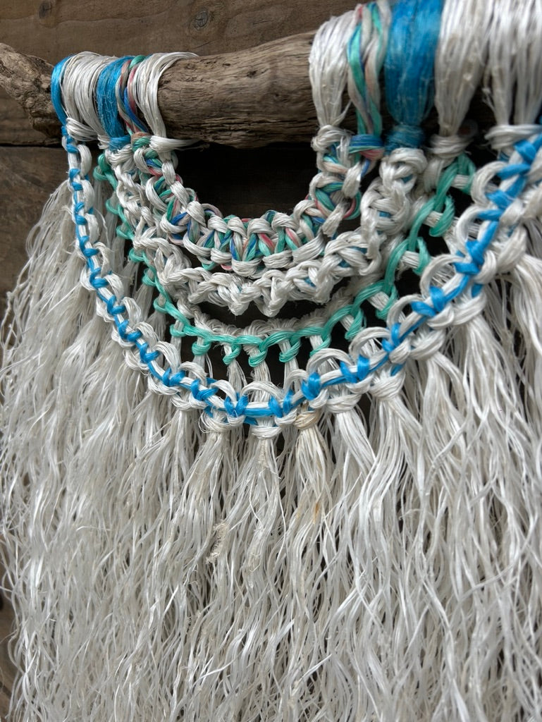 Detail shot of coastal wall hanging, with sea blue and green knots and a weathered white fringe, handcrafted from ocean waste, set on driftwood.