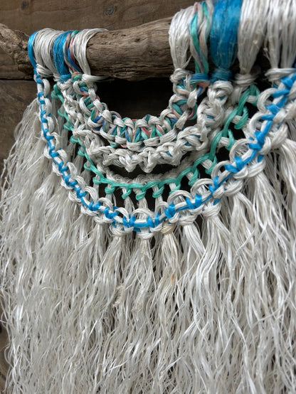 Detail shot of coastal wall hanging, with sea blue and green knots and a weathered white fringe, handcrafted from ocean waste, set on driftwood.