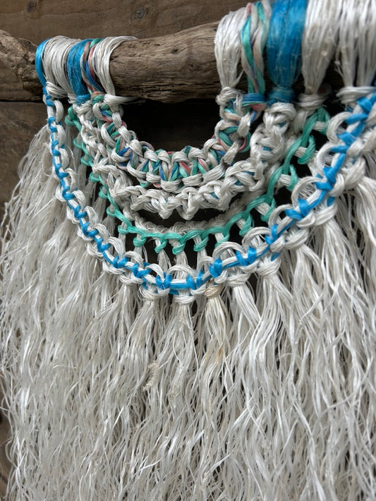 Detail shot of coastal wall hanging, with sea blue and green knots and a weathered white fringe, handcrafted from ocean waste, set on driftwood.