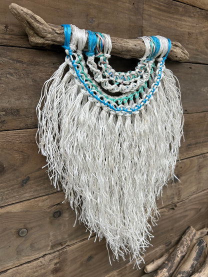 Side shot of handmade, textured, reclaimed fishing rope macramé wall hanging in weathered white with sea greens and blues, set on driftwood.