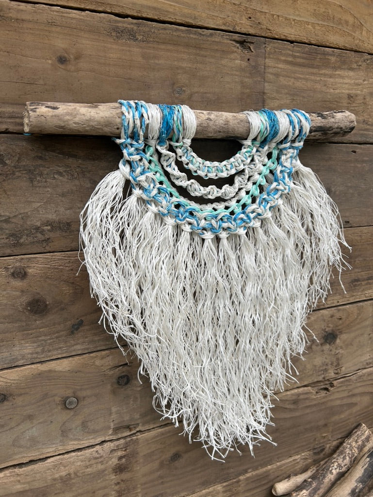 Side shot of handmade, sustainable fishing rope wall art, with a weathered white fringe and sea blues and greens, set on driftwood.