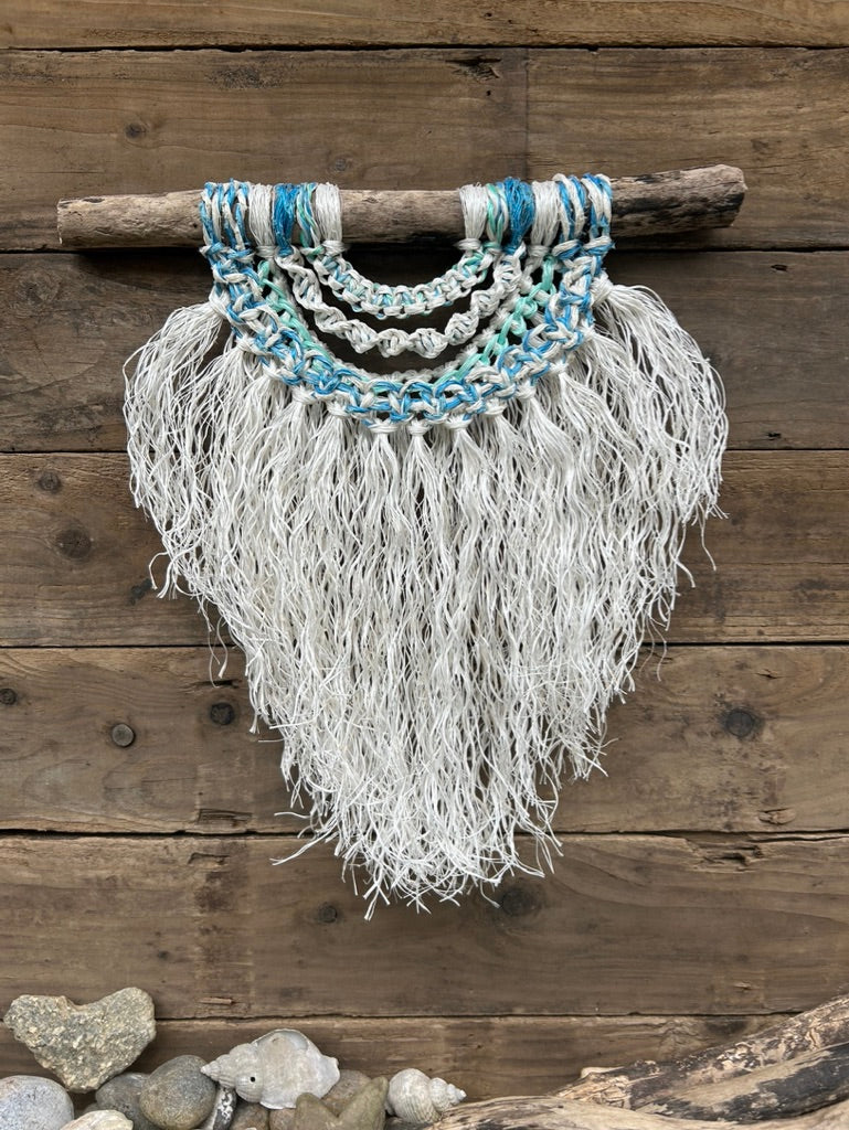 Textured, multi-layered wall hanging in soft white with sea blues and greens, handcrafted sustainably from reclaimed fishing ropes, set on driftwood.