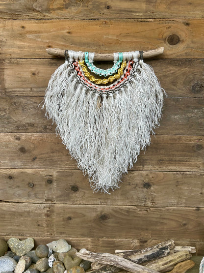 Textured, multi-layered macramé wall hanging, handcrafted from repurposed fishing ropes, with a weathered white fringe and reggae inspired tones of green, yellow, red and black, set on driftwood.