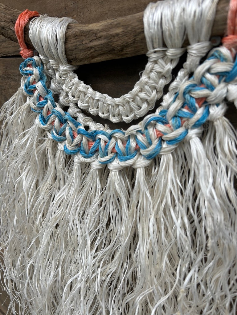 Detail photo of a handcrafted macramé wall hanging made from ocean waste, with a soft white fringe, blue and white knots and a touch of sun-faded orange, set on driftwood.