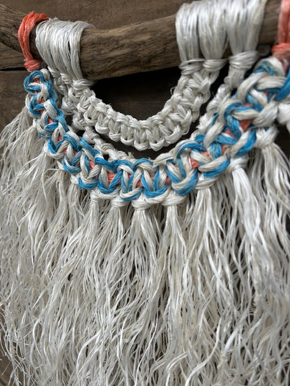 Detail photo of a handcrafted macramé wall hanging made from ocean waste, with a soft white fringe, blue and white knots and a touch of sun-faded orange, set on driftwood.
