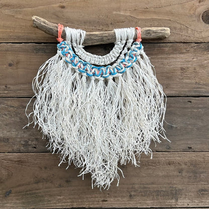 Textured coastal interior wall art, hand-knotted from reclaimed fishing ropes, with a soft white fringe and blue and white knots and a touch of sun-bleached orange, set on driftwood.