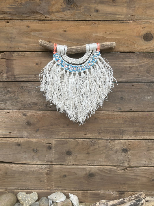 Coastal decor rope wall art, handmade by recycling retired fishing ropes, with a soft white fringe, white and blue knots and a touch of sun-faded orange, set on driftwood.