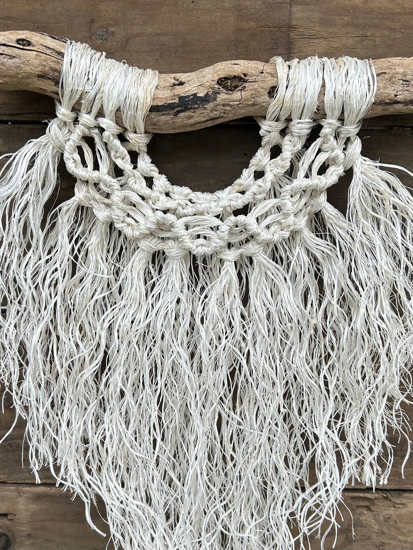 Close-up of eco-friendly, ocean inspired wall art, handcrafted from retired fishing rope, with a textured soft white fringe and wave knot band, set on driftwood.